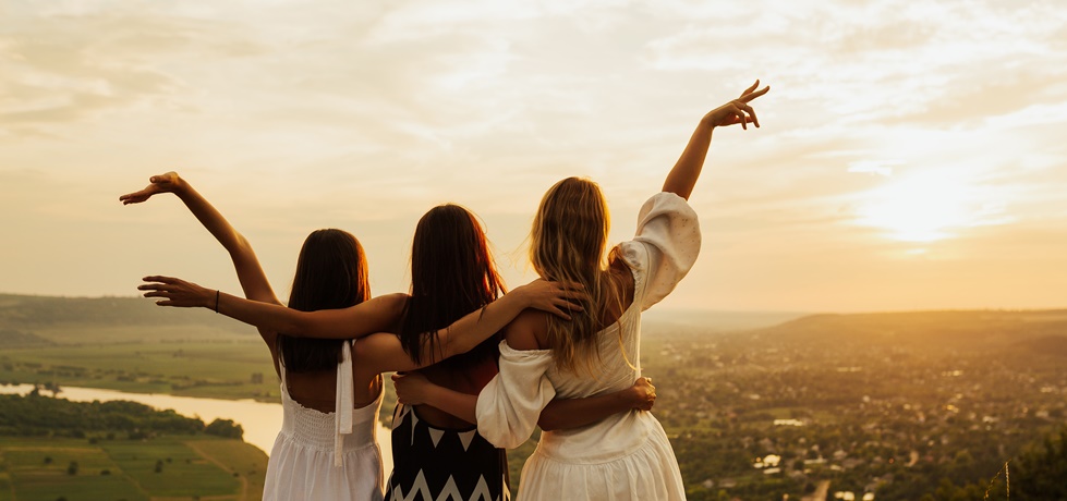 Três mulheres abraçadas ao pôr do sol, simbolizando amizade e conexão no grupo Amizade Feminina da comunidade Tribou.